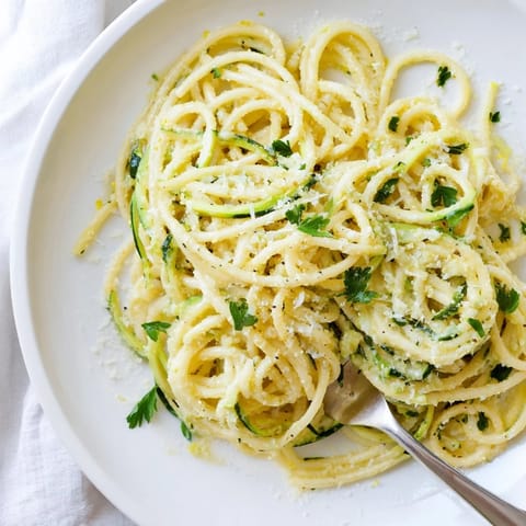 Plated Lemon Zucchini Pasta topped with grated Parmesan and parsley, steaming beside a wine glass on a rustic wooden table.