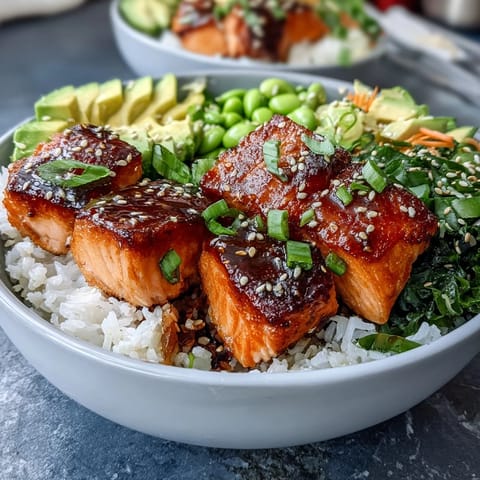Golden brown broiled salmon cubes rest on fluffy white rice beside crisp cucumber slices, shredded carrots, and bright green edamame in a healthy Baked Salmon Rice Bowl.